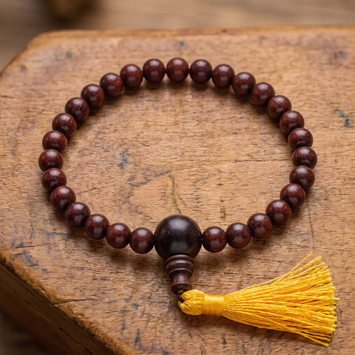 Draufsicht auf das Power-Bracelet Rosenholz auf altem Holz: Reihe aus dunklen Rosenholzperlen mit größerer Guruperle und gelber Quaste