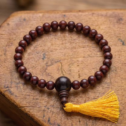 Draufsicht auf das Power-Bracelet Rosenholz auf altem Holz: Reihe aus dunklen Rosenholzperlen mit größerer Guruperle und gelber Quaste