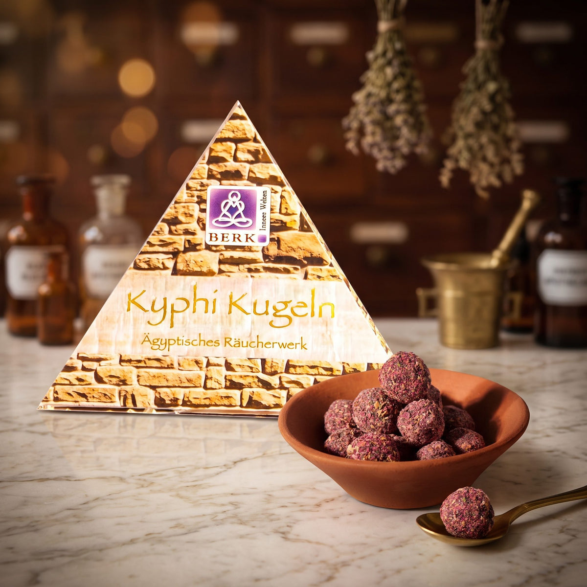 Kyphi Kugeln von Berk: die Pyramidenverpackung auf der Marmortheke einer alten Apotheke am Abend, daneben eine Terracottaschale mit rosafarbenen Kyphi-Kugeln und ein Messinglöffel – Spirit of Gaia