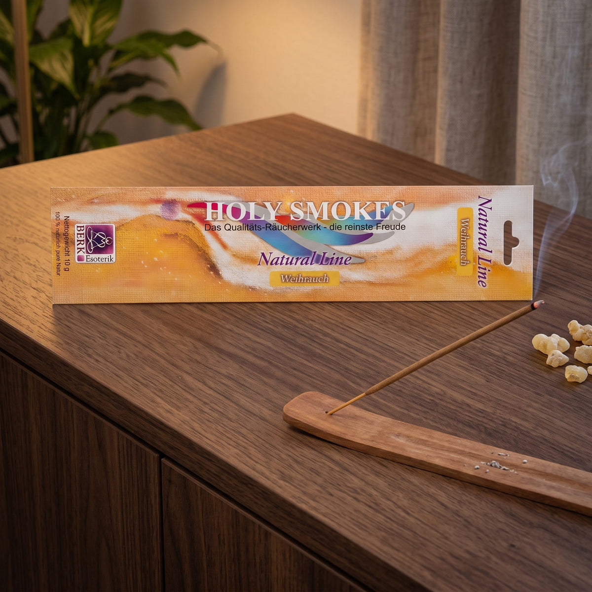 Weihrauch Räucherstäbchen Natural Line: glimmendes Stäbchen im Holzhalter auf einem dunklen Sideboard, dahinter die Packung, am Abend