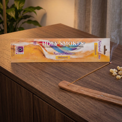 Weihrauch Räucherstäbchen Natural Line: glimmendes Stäbchen im Holzhalter auf einem dunklen Sideboard, dahinter die Packung, am Abend