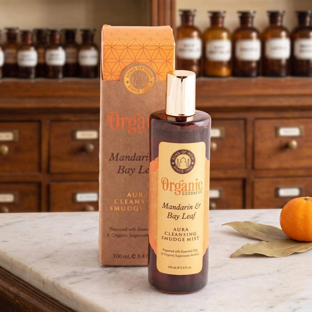 Aura Reinigungsspray Mandarine und Lorbeerblatt von Organic Goodness: braune Sprühflasche mit goldener Kappe neben der Kraftpapier-Schachtel auf einer Marmortheke vor einem Apothekerschrank