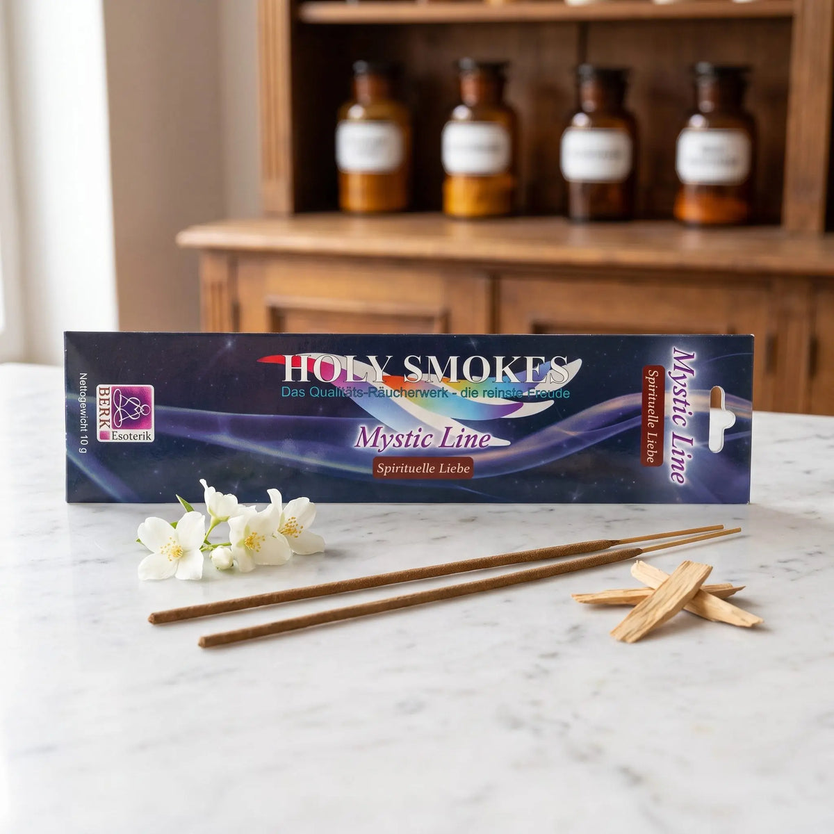 Spirituelle Liebe Räucherstäbchen Holy Smokes Mystic Line: Packung auf einer Marmortheke vor einem Apothekerschrank, davor zwei lose Räucherstäbchen, Jasminblüten und Sandelholz – Spirit of Gaia