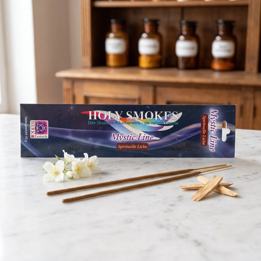 Spirituelle Liebe Räucherstäbchen Holy Smokes Mystic Line: Packung auf einer Marmortheke vor einem Apothekerschrank, davor zwei lose Räucherstäbchen, Jasminblüten und Sandelholz – Spirit of Gaia