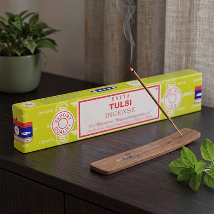 Satya Tulsi Räucherstäbchen: glimmendes Räucherstäbchen mit feinem Rauch im Holzhalter, dahinter die Packung, auf einem Sideboard am Abend