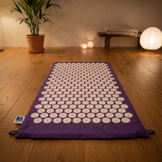 Akupressurmatte Lebensblume, 78 × 45 cm, Baumwollbezug lila, weiße Akupressurspitzen: ausgelegt auf dem Holzboden eines ruhigen Meditationsraums am Abend – Spirit of Gaia