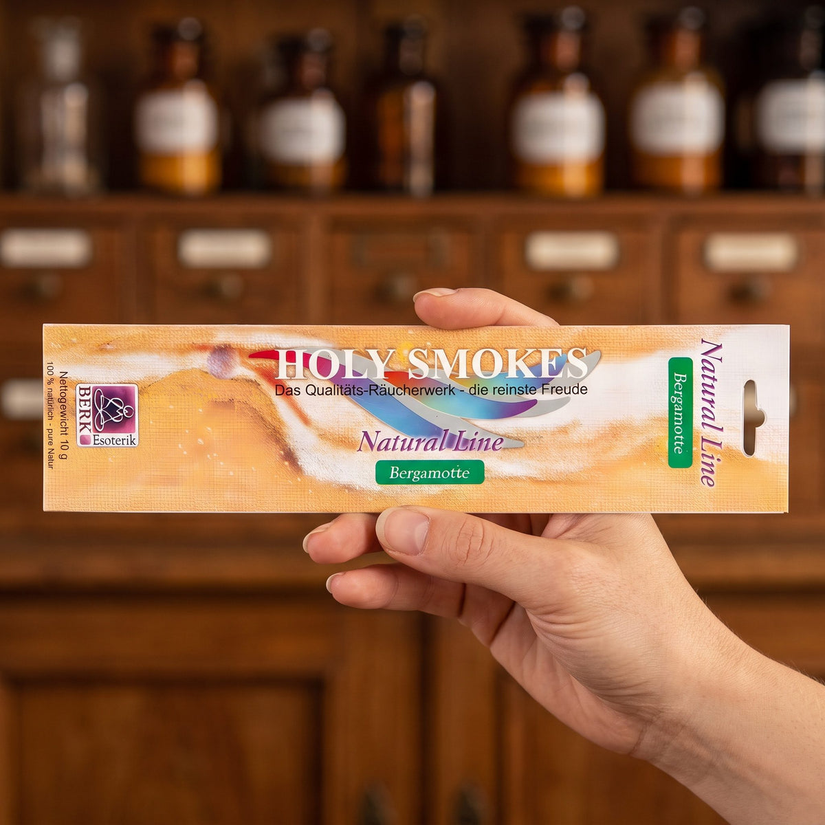 Eine Hand hält die flache Packung Bergamotte Natural Line von Holy Smokes, etwa 24 cm lang, vor einem Holzschrank mit Schubladen