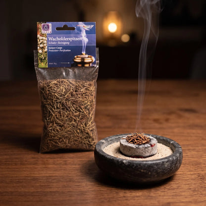 Wacholderspitzen, sortenreines Räucherwerk von Berk, 50 g: eine Prise Wacholderspitzen glimmt auf der Räucherkohle, dahinter die Tüte, feiner Rauch steigt auf einer dunklen Holzplatte am Abend auf – Spirit of Gaia