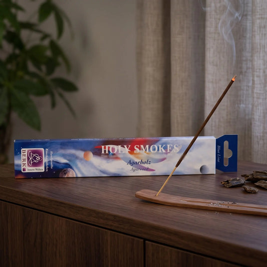 Agarholz Räucherstäbchen Holy Smokes Blue Line: glimmendes Räucherstäbchen mit feinem Rauch im Holzhalter, dahinter die Packung, auf einem Sideboard am Abend