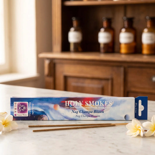 Nag Champa Blüten Räucherstäbchen Blue Line: Packung auf einer Marmortheke vor einem Apothekerschrank, daneben zwei lose Räucherstäbchen und ein paar cremeweiße Champablüten – Spirit of Gaia