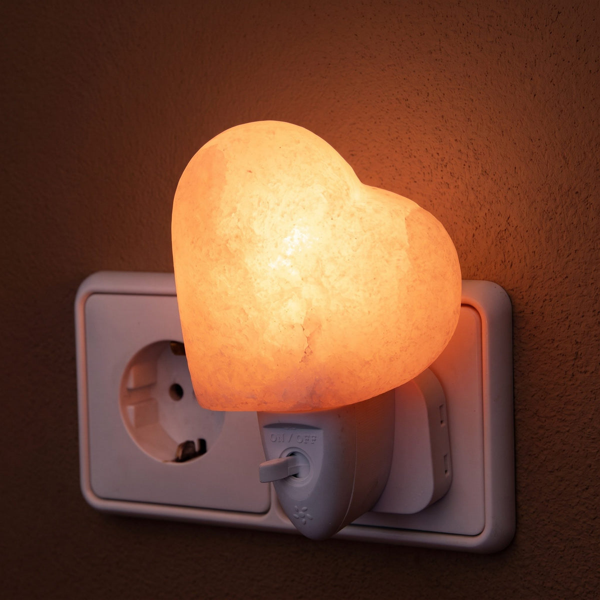 Salzkristall-Nachtlicht Herz in einer Steckdose leuchtet warm rosaorange vor einer dunklen Putzwand