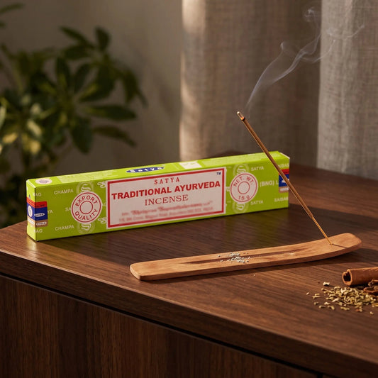 Satya Traditional Ayurveda Räucherstäbchen: glimmendes Räucherstäbchen mit feinem Rauch im Holzhalter, dahinter die Packung, auf einem Sideboard am Abend