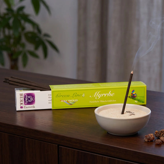 Myrrhe Räucherstäbchen naturrein, Holy Smokes Green Line: glimmendes Räucherstäbchen in einer Keramikschale mit Sand, dahinter die geöffnete Packung, auf einem Sideboard am Abend