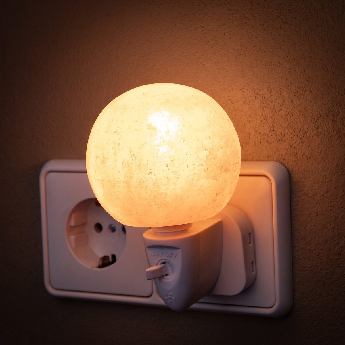 Salzkristall-Nachtlicht Kugel in einer Steckdose leuchtet warm vor einer dunklen Putzwand