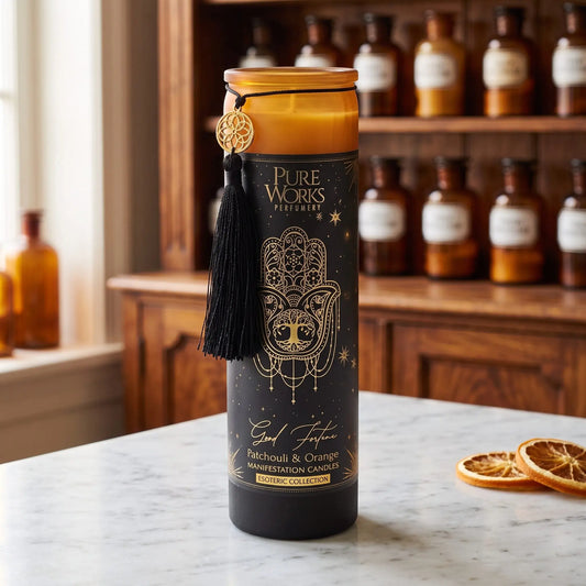 Manifestationskerze Good Fortune (Patchouli & Orange) von Pure Works: hohe Glaskerze mit orangefarbenem Glasrand, schwarzer Quaste und Goldanhänger auf Marmortheke vor Apothekerschrank, daneben getrocknete Orangenscheiben – Spirit of Gaia