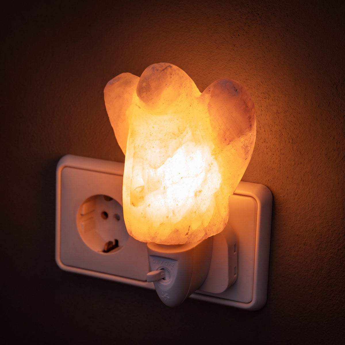 Salzkristall-Nachtlicht Engel in einer Steckdose leuchtet warm vor einer dunklen Putzwand