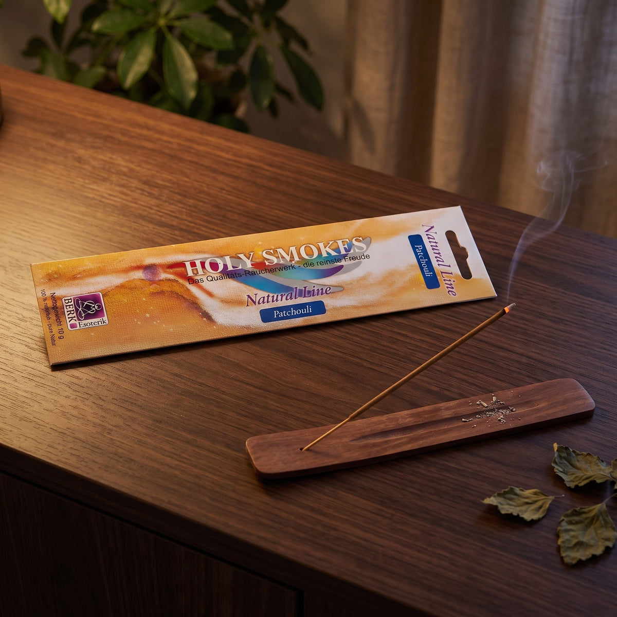 Patchouli Räucherstäbchen Natural Line: glimmendes Stäbchen im Holzhalter auf einem dunklen Sideboard, dahinter die Packung, am Abend
