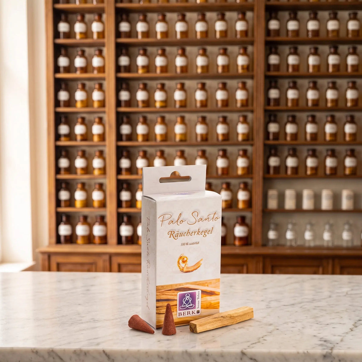 Räucherpyramiden Palo Santo von Berk: Schachtel auf einer Marmortheke vor einem Apothekerschrank, davor zwei lose Räucherkegel und ein Stück Palo-Santo-Holz – Spirit of Gaia