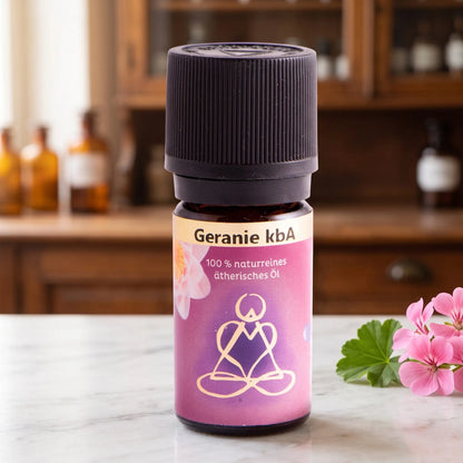 Ätherisches Öl Geranie: Fläschchen aus Braunglas auf Marmortheke vor Apothekerschrank, daneben Geranienblüten – Spirit of Gaia