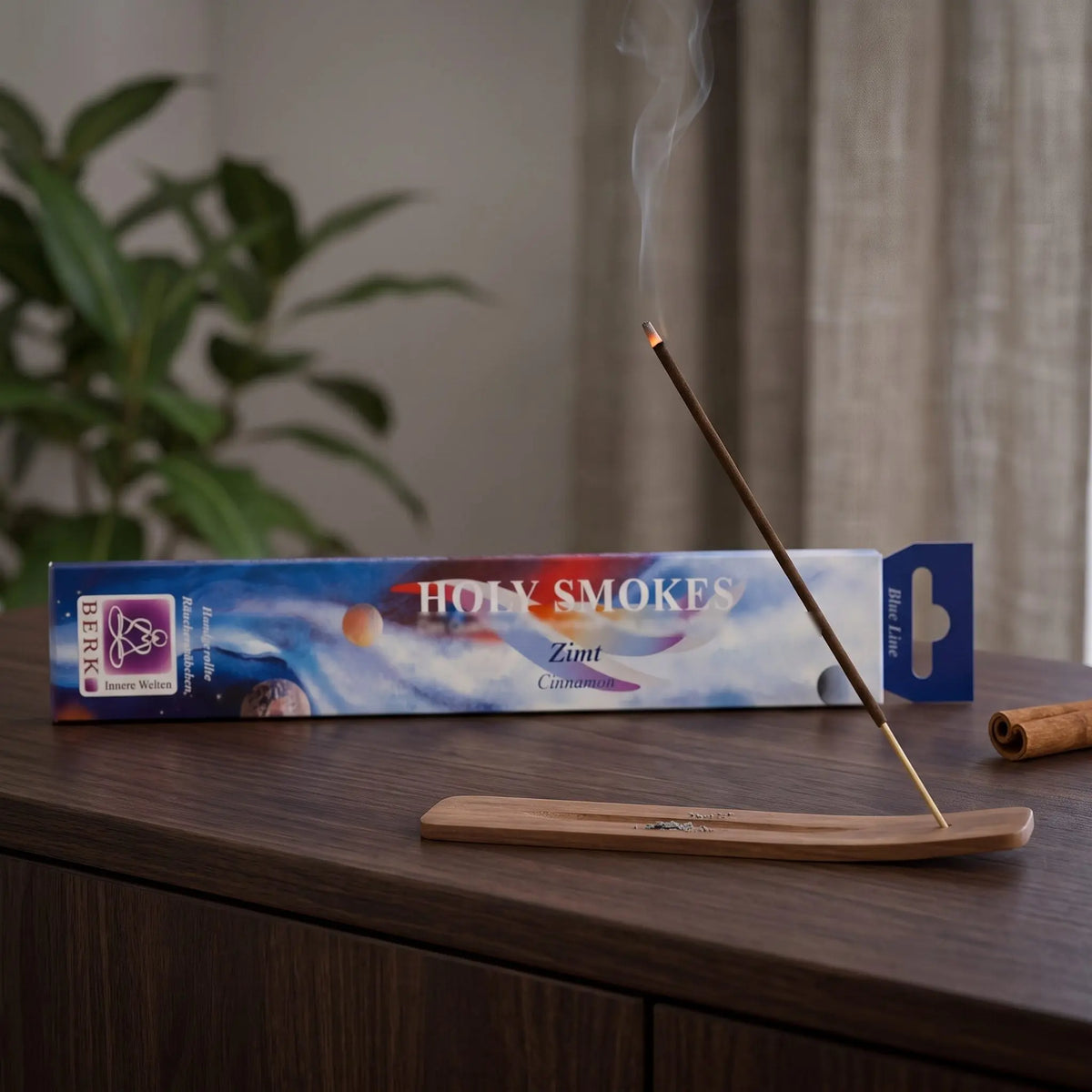 Zimt Räucherstäbchen Holy Smokes Blue Line: glimmendes Räucherstäbchen mit feinem Rauch im Holzhalter, dahinter die Packung, auf einem Sideboard am Abend
