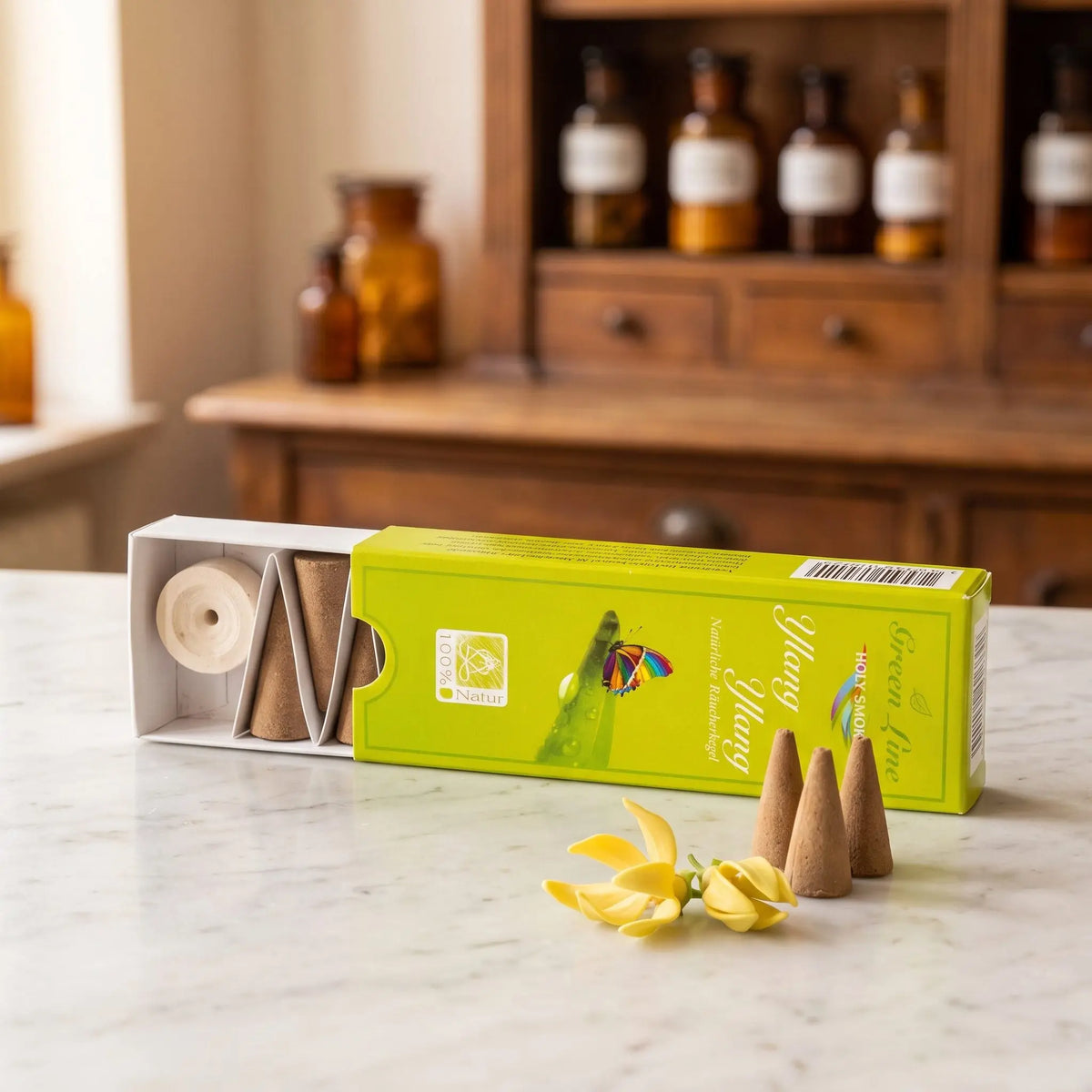 Ylang Ylang Räucherkegel naturrein, Holy Smokes Green Line: geöffnete Packung auf einer Marmortheke vor einem Apothekerschrank, davor lose Räucherkegel und Ylang-Ylang-Blüten – Spirit of Gaia