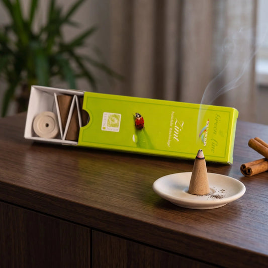 Zimt Räucherkegel naturrein, Holy Smokes Green Line: glimmender Räucherkegel mit feinem Rauch auf einem Keramiktellerchen, dahinter die geöffnete Packung, auf einem Sideboard am Abend