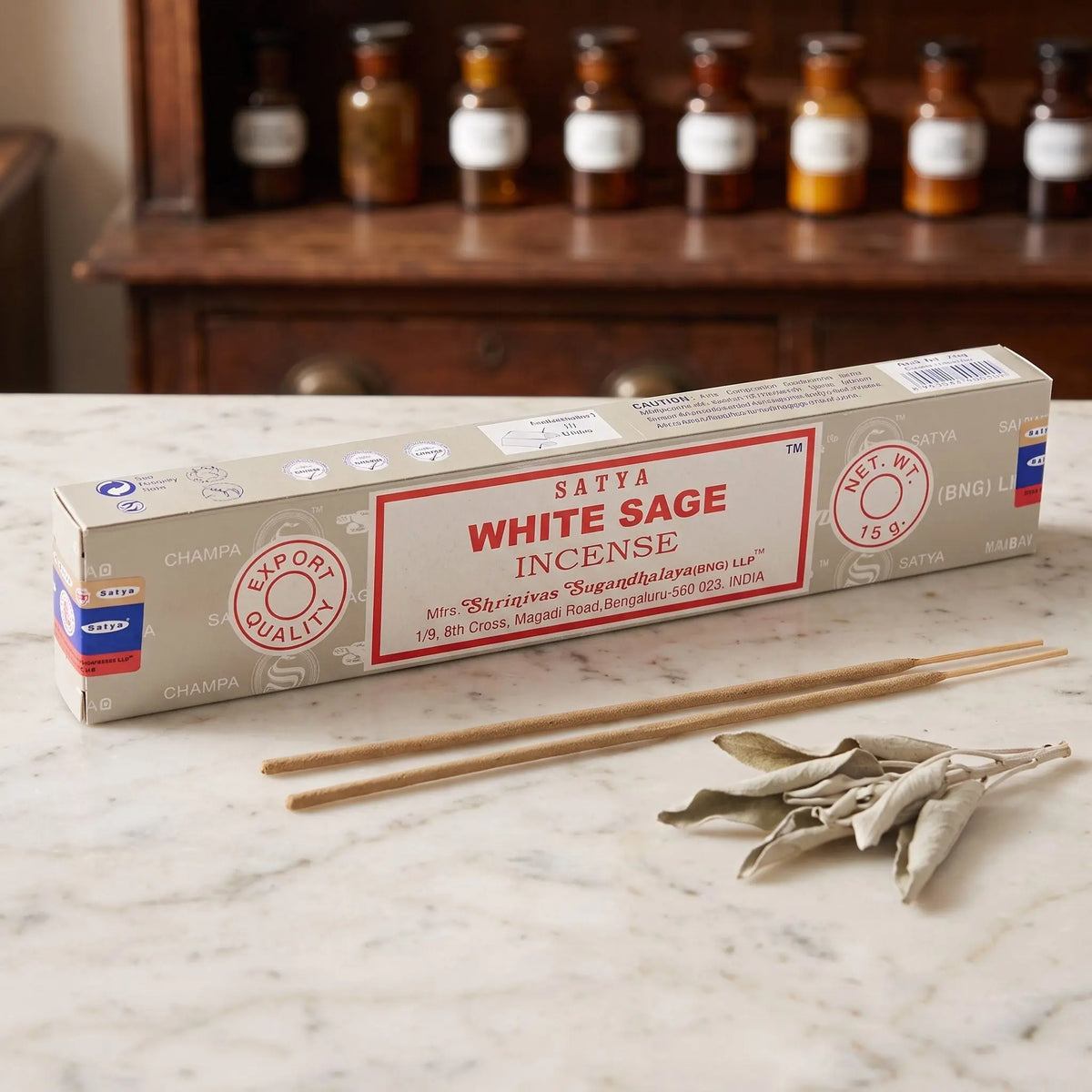 Satya White Sage Räucherstäbchen: 15-g-Packung auf einer Marmortheke vor einem Apothekerschrank, daneben zwei lose Räucherstäbchen – Spirit of Gaia