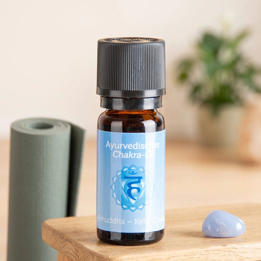 Ayurvedisches Chakra-Öl Vishuddha (Kehl-Chakra): 10-ml-Fläschchen auf einem Holzhocker neben einer gerollten Yogamatte, daneben blauer Chalcedon