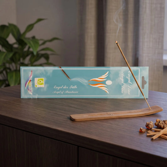 Engel der Fülle Räucherstäbchen Holy Smokes: glimmendes Räucherstäbchen mit feinem Rauch im Holzhalter, dahinter die Packung, auf einem Sideboard am Abend