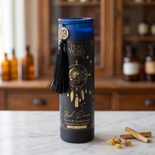 Manifestationskerze Boho Dreams (Myrrhe & Sandelholz) von Pure Works: hohe Glaskerze mit blauem Glasrand, schwarzer Quaste und Goldanhänger auf Marmortheke vor Apothekerschrank, daneben Myrrheharz und Sandelholzspäne – Spirit of Gaia