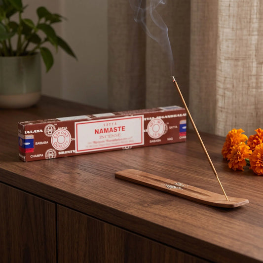 Satya Namaste Räucherstäbchen: glimmendes Räucherstäbchen mit feinem Rauch im Holzhalter, dahinter die Packung, auf einem Sideboard am Abend