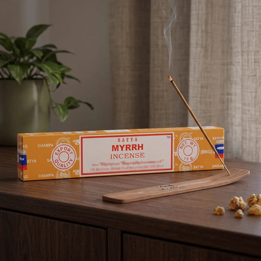 Satya Myrrh Räucherstäbchen: glimmendes Räucherstäbchen mit feinem Rauch im Holzhalter, dahinter die Packung, auf einem Sideboard am Abend