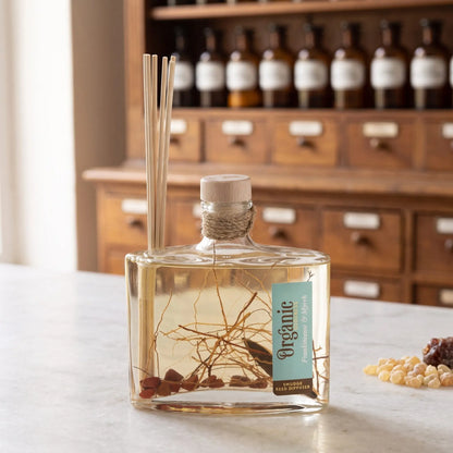 Reed Diffuser Weihrauch und Myrrhe von Organic Goodness: eckige Glasflasche mit Holzstopfen, Jutekordel und Schilfrohrstäbchen auf einer Marmortheke vor einem Apothekerschrank