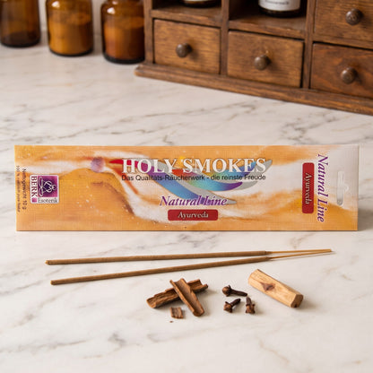 Ayurveda Räucherstäbchen der Holy Smokes Natural Line von Berk: flache Packung auf einer Marmortheke vor einem alten Holzschrank mit Schubladen und Bernsteinglasflaschen, davor zwei lose Stäbchen und Zimtrinde, Nelken und ein Stück Sandelholz