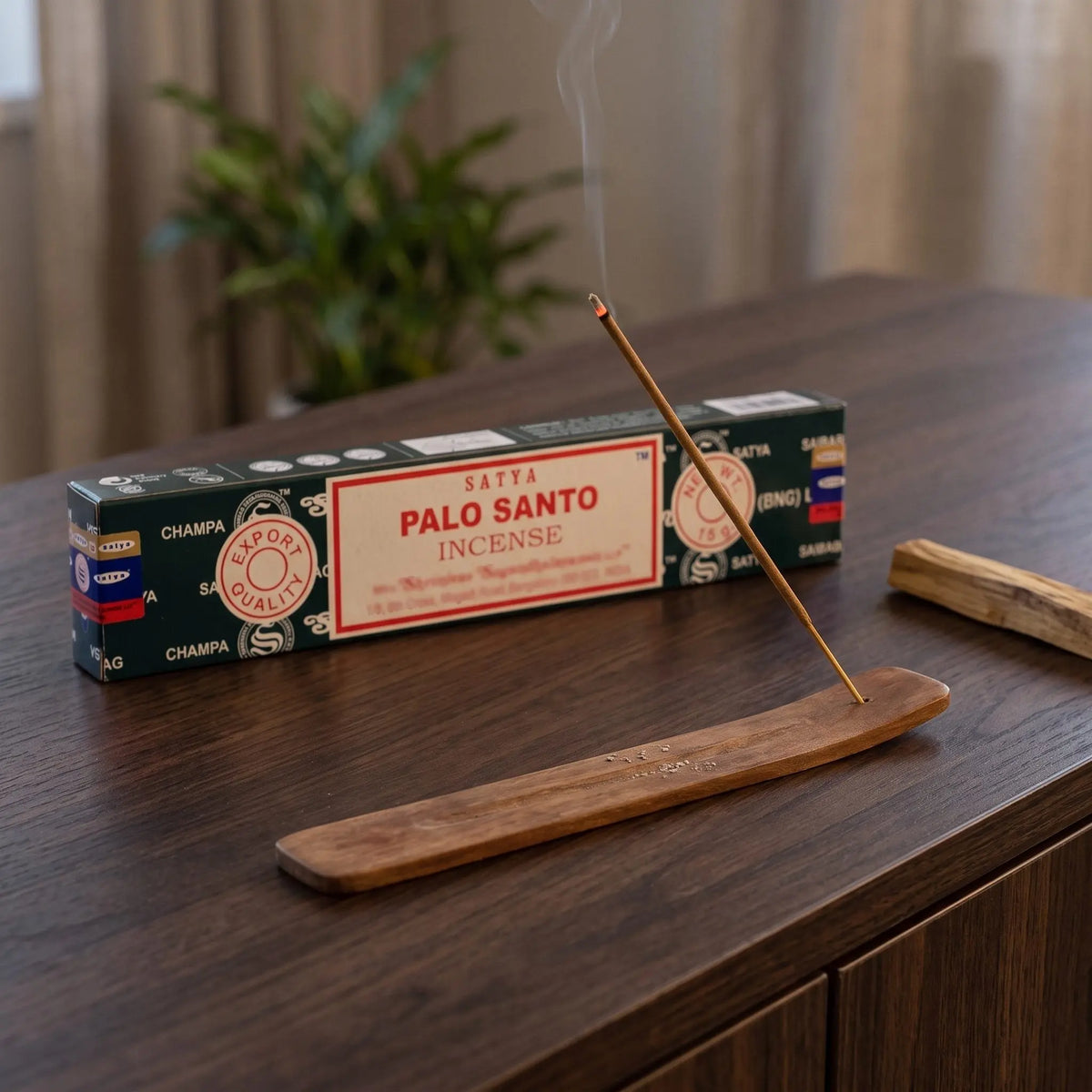Satya Palo Santo Räucherstäbchen: glimmendes Räucherstäbchen mit feinem Rauch im Holzhalter, dahinter die Packung, auf einem Sideboard am Abend