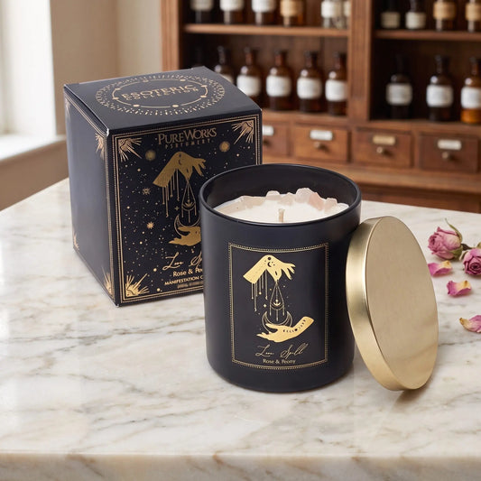 Manifestationskerze Love Spell mit Rosenquarz (Rose & Pfingstrose) von Pure Works: große Kerze im schwarzen Glas mit Golddeckel und schwarze Schachtel mit Goldprägung auf Marmortheke vor Apothekerschrank, daneben getrocknete Rosenknospen – Spirit of Gaia