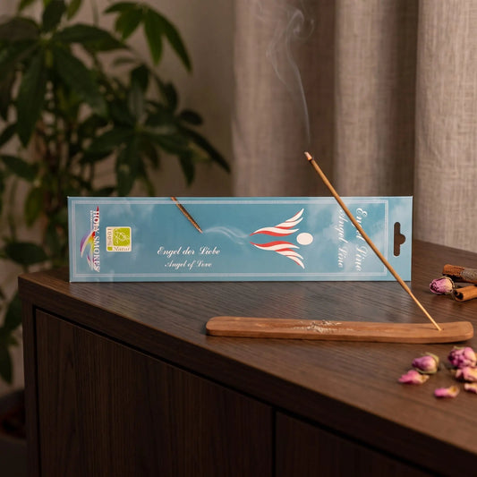 Engel der Liebe Räucherstäbchen Holy Smokes: glimmendes Räucherstäbchen mit feinem Rauch im Holzhalter, dahinter die Packung, auf einem Sideboard am Abend
