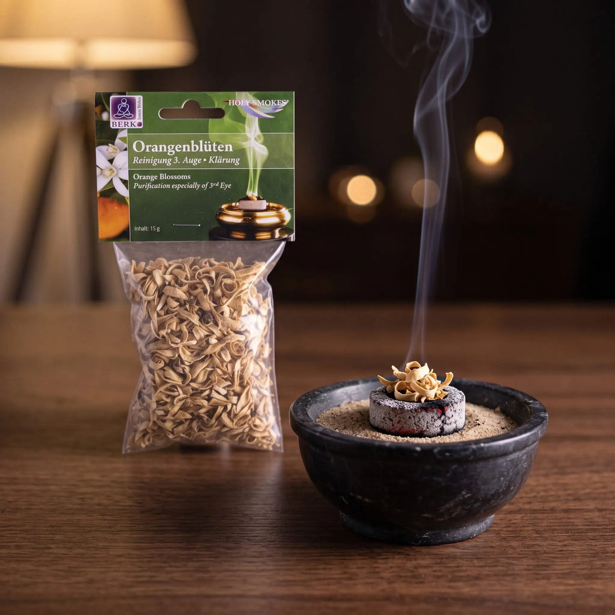 Orangenblüten, sortenreines Räucherwerk von Berk, 15 g: eine Prise Orangenblüten glimmt auf der Räucherkohle, dahinter die Tüte, feiner Rauch steigt auf einer dunklen Holzplatte am Abend auf – Spirit of Gaia