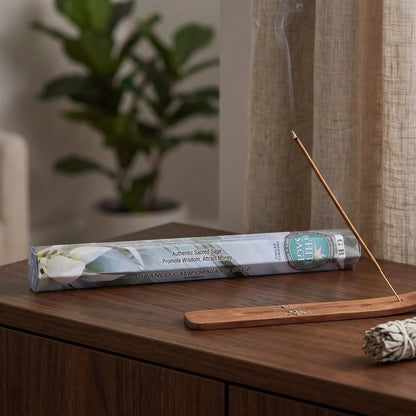 GR White Sage Räucherstäbchen: glimmendes Räucherstäbchen mit feinem Rauch im Holzhalter, dahinter die Sechseck-Packung, auf einem Sideboard am Abend