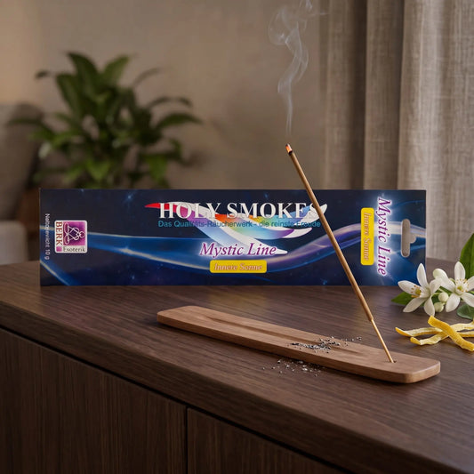 Innere Sonne Räucherstäbchen Holy Smokes Mystic Line: glimmendes Räucherstäbchen mit feinem Rauch im Holzhalter, dahinter die Packung, auf einem Sideboard am Abend