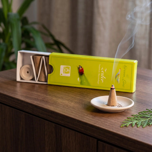 Zeder Räucherkegel naturrein, Holy Smokes Green Line: glimmender Räucherkegel mit feinem Rauch auf einem Keramiktellerchen, dahinter die geöffnete Packung, auf einem Sideboard am Abend