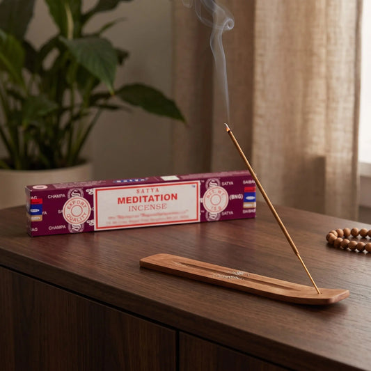 Satya Meditation Räucherstäbchen: glimmendes Räucherstäbchen mit feinem Rauch im Holzhalter, dahinter die Packung, auf einem Sideboard am Abend