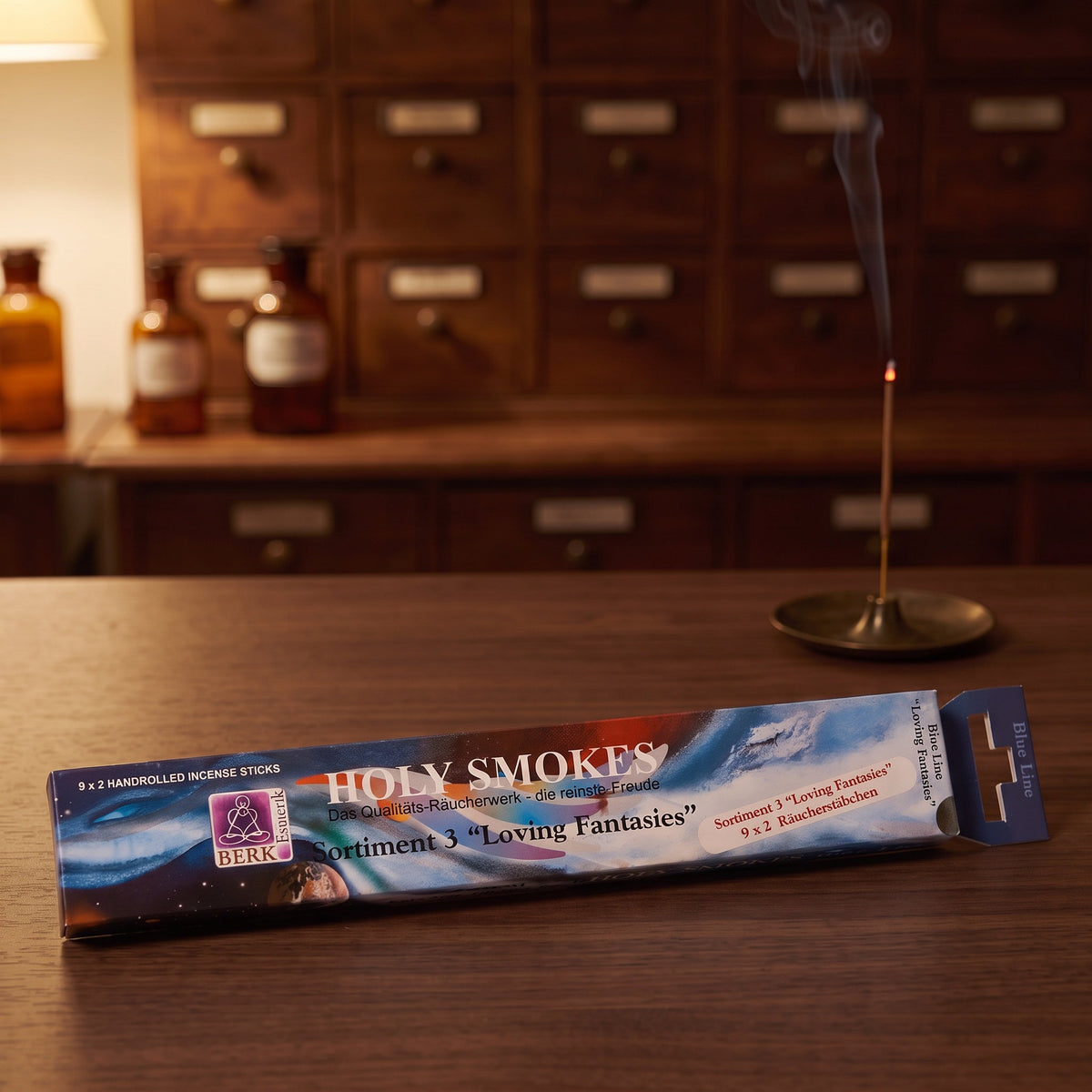 Packung Loving Fantasies Sortiment 3 am Abend auf einem Sideboard, dahinter glimmt ein Räucherstäbchen in einem Messinghalter