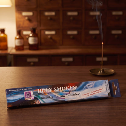Packung Loving Fantasies Sortiment 3 am Abend auf einem Sideboard, dahinter glimmt ein Räucherstäbchen in einem Messinghalter
