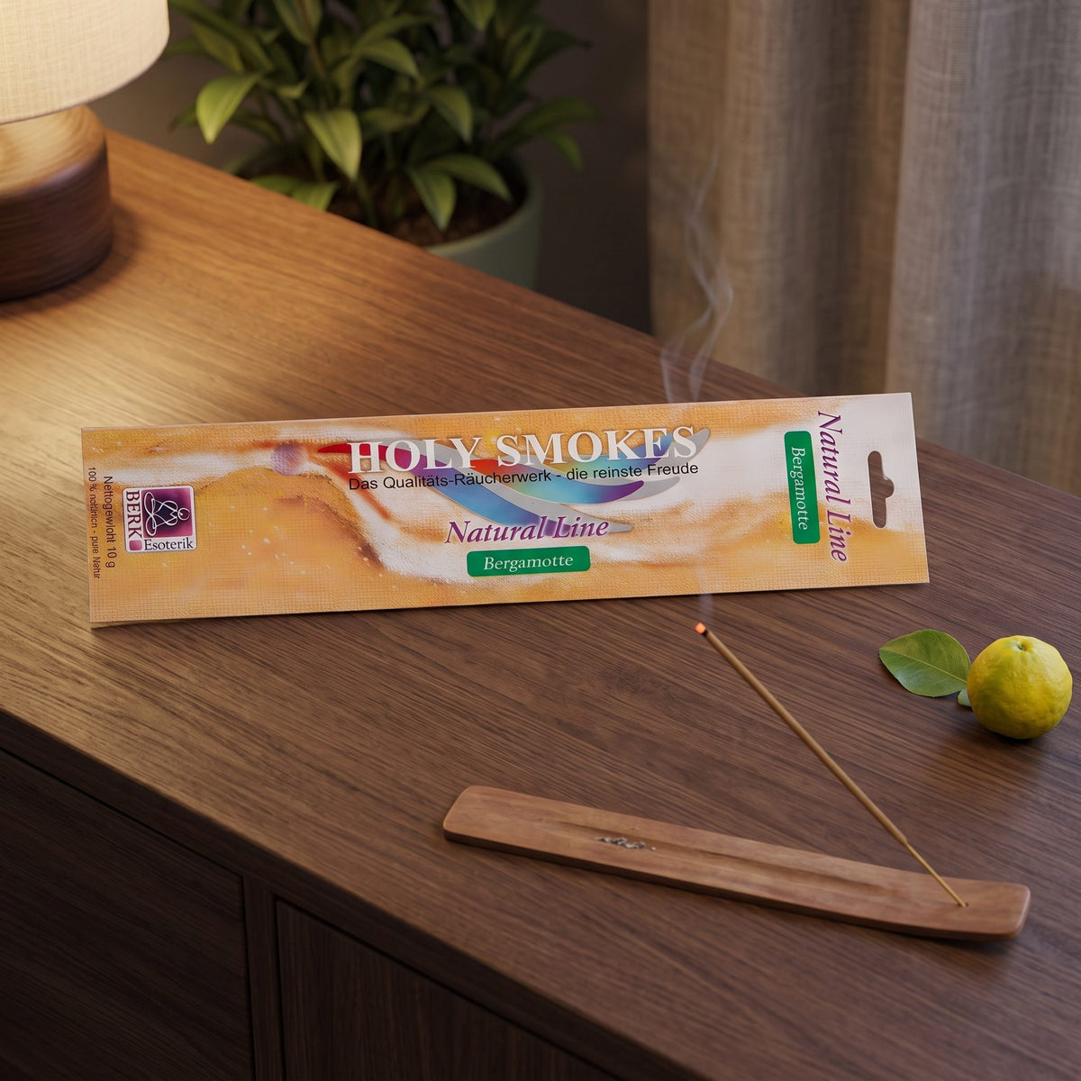 Bergamotte Räucherstäbchen Natural Line: glimmendes Stäbchen im Holzhalter auf einem dunklen Sideboard, dahinter die Packung, am Abend
