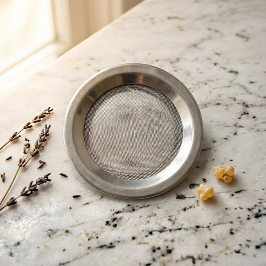 Räuchersieb aus Edelstahl: flacher Ring mit hochgezogenem Rand und feinmaschigem Drahtgewebe – auf der Marmortheke vor dem Apothekerschrank