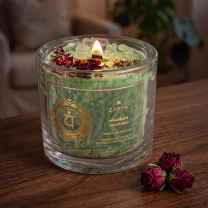 Chakra-Kerze Herzchakra Anahata mit Fluorit und Rosenquarz, Duft Rose und Mandel: brennende Kerze mit kleiner Flamme am Abend auf einer dunklen Holzplatte im Wohnzimmer, daneben getrocknete Rosenknospen – Spirit of Gaia
