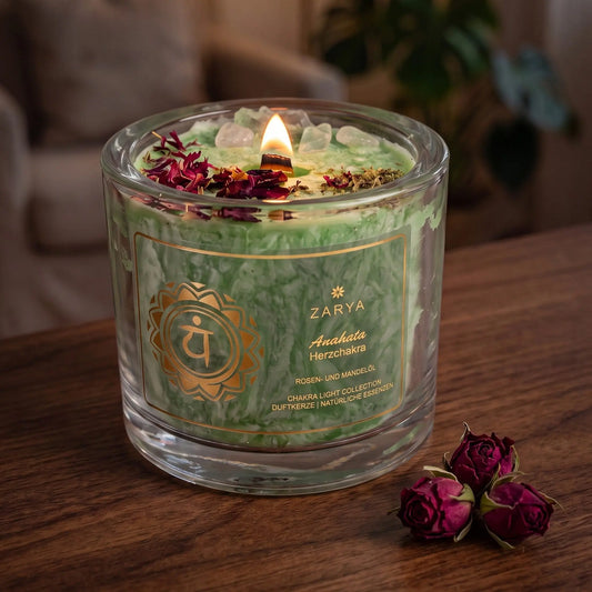 Chakra-Kerze Herzchakra Anahata mit Fluorit und Rosenquarz, Duft Rose und Mandel: brennende Kerze mit kleiner Flamme am Abend auf einer dunklen Holzplatte im Wohnzimmer, daneben getrocknete Rosenknospen – Spirit of Gaia