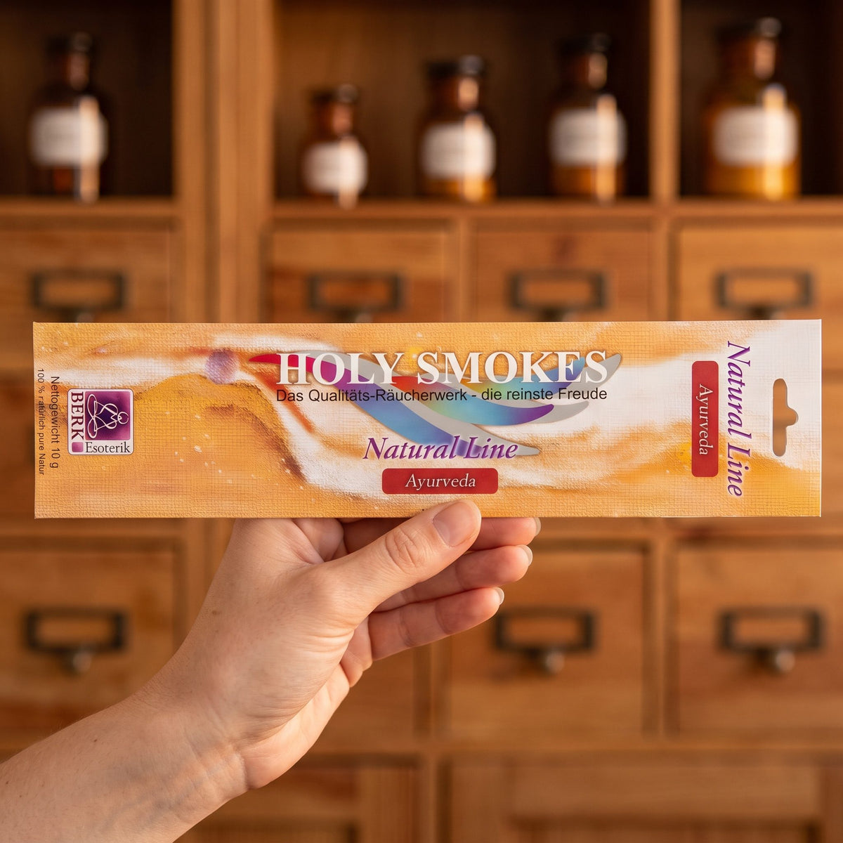 Eine Hand hält die flache Packung Ayurveda Natural Line von Holy Smokes, etwa 24 cm lang, vor einem Holzschrank mit Schubladen