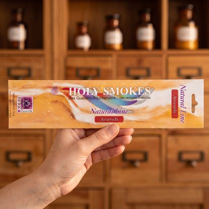 Eine Hand hält die flache Packung Ayurveda Natural Line von Holy Smokes, etwa 24 cm lang, vor einem Holzschrank mit Schubladen